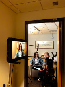 A young white woman sits in a chair while an older white man reviews a script with her for a video shoot in which she plays a student in need of counseling. The photo is framed through an open doorway with a view of the camera screen in the foreground.