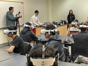 A classroom with multiple students sitting at desks with virtual reality headsets on.
