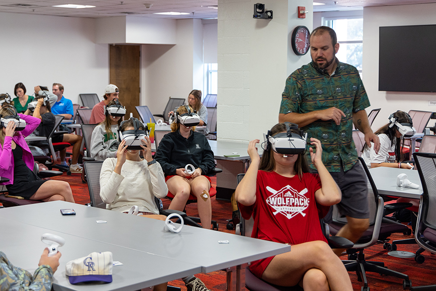A group of students in a classroom wearing virtual reality headsets while a male professor looks on. 