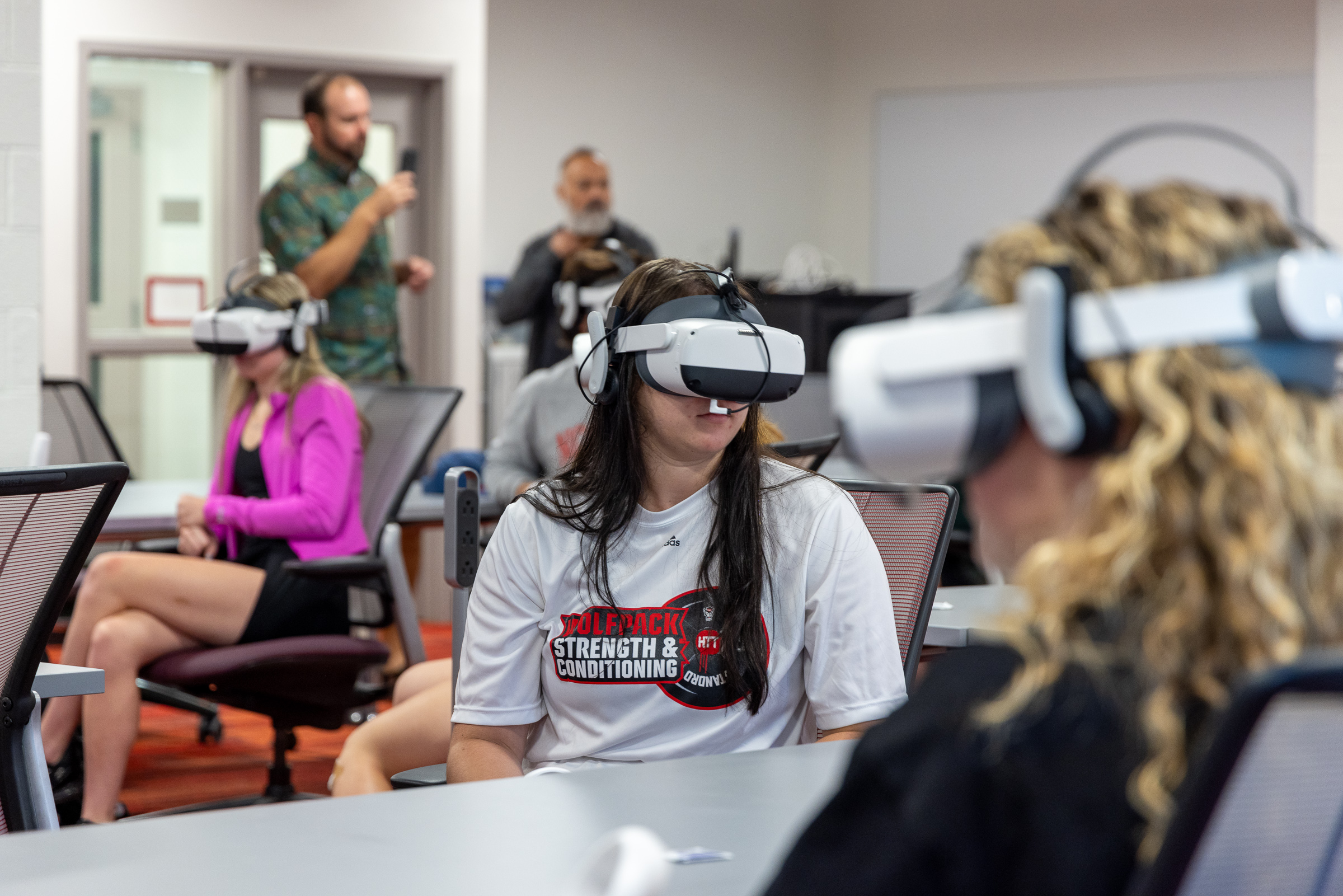 A white student with long brown hair uses a virtual reality headset while sitting at a desk in a classroom.