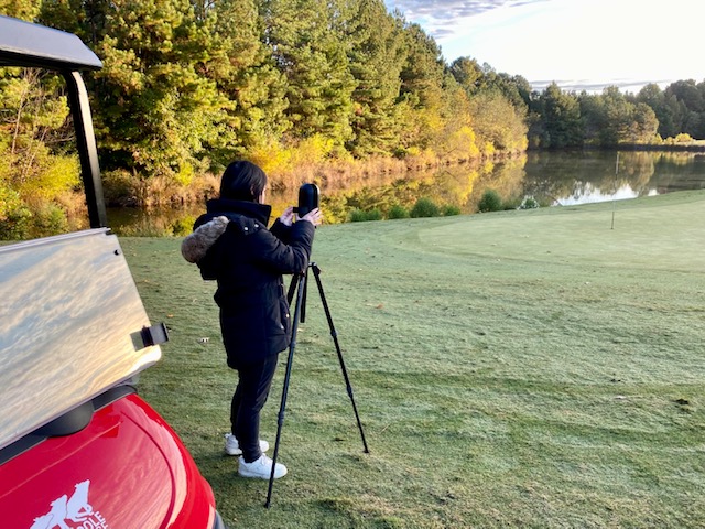 A person standing in a grassy field using a camera on a tripod.